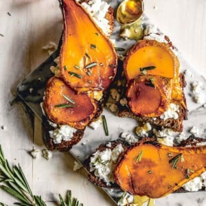 Roasted pear with honey and rosemary on sourdough, on a marble slab tray, beside a jar of honey, a spoon, and sprigs of rosemary.