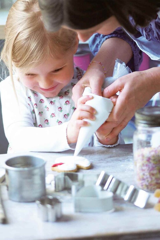 Royal icing being piped onto a cookie by a young girl being helped by her mother, surrounded by jars of sprinkles, cookie cutters, and wooden spoons.