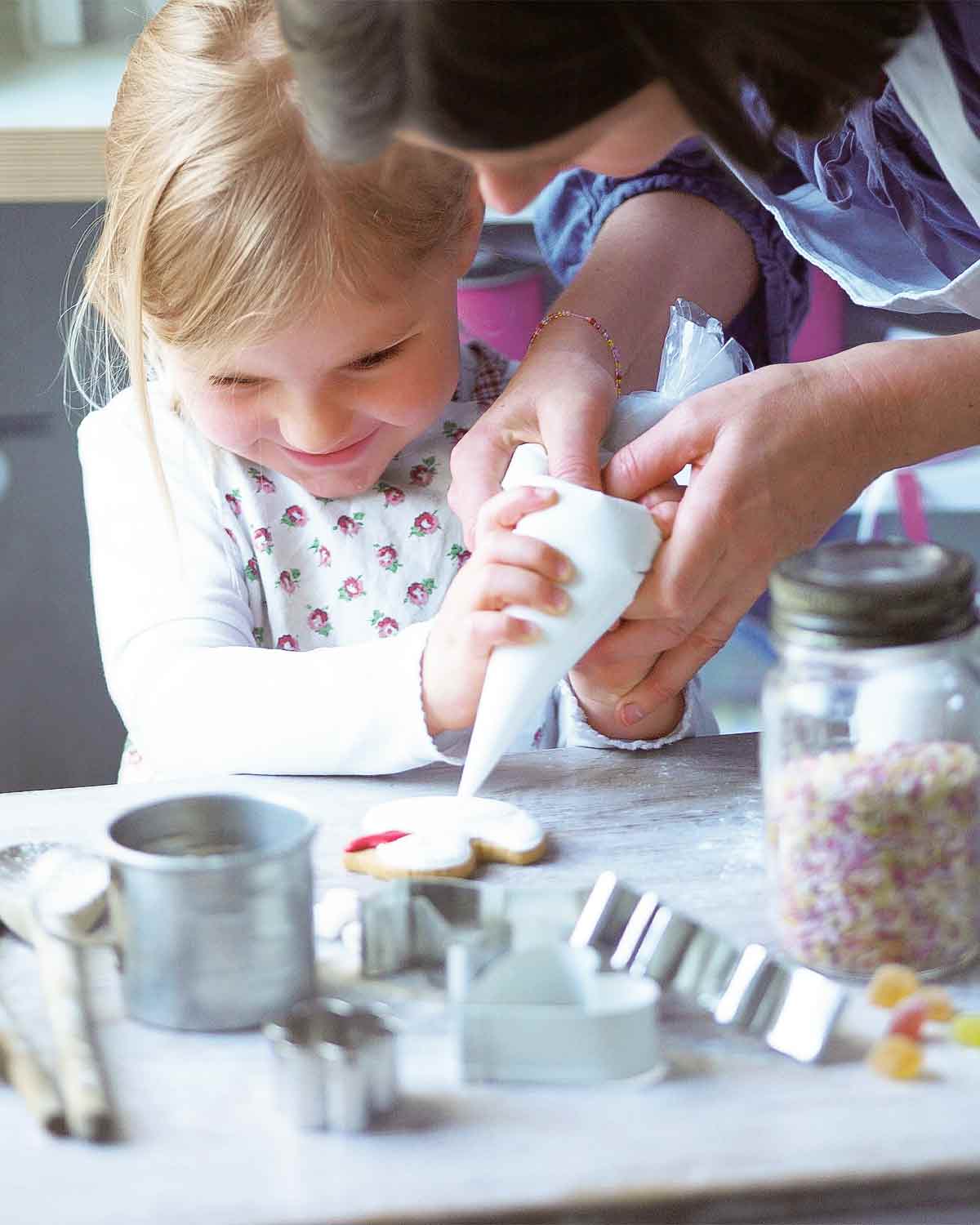 Royal icing being piped onto a cookie by a young girl being helped by her mother, surrounded by jars of sprinkles, cookie cutters, and wooden spoons.