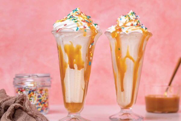 Salted caramel funfetti milkshakes in tall parfait glasses with sprinkles, beside a jar of sprinkles, a linen napkin, and a jar of caramel sauce.