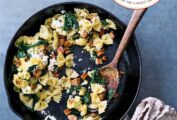 Skillet cauliflower pasta with farfalle, andouille sausage, spinach, and goat cheese in a cast iron pan, a wooden spoon and striped towel nearby.