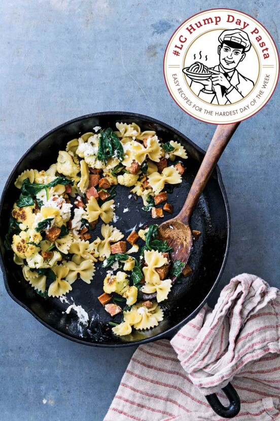 Skillet cauliflower pasta with farfalle, andouille sausage, spinach, and goat cheese in a cast iron pan, a wooden spoon and striped towel nearby.