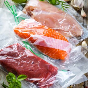 Vacuum sealed pieces of salmon, chicken, and steak on a wooden table with basil sprigs and garlic cloves scattered around..