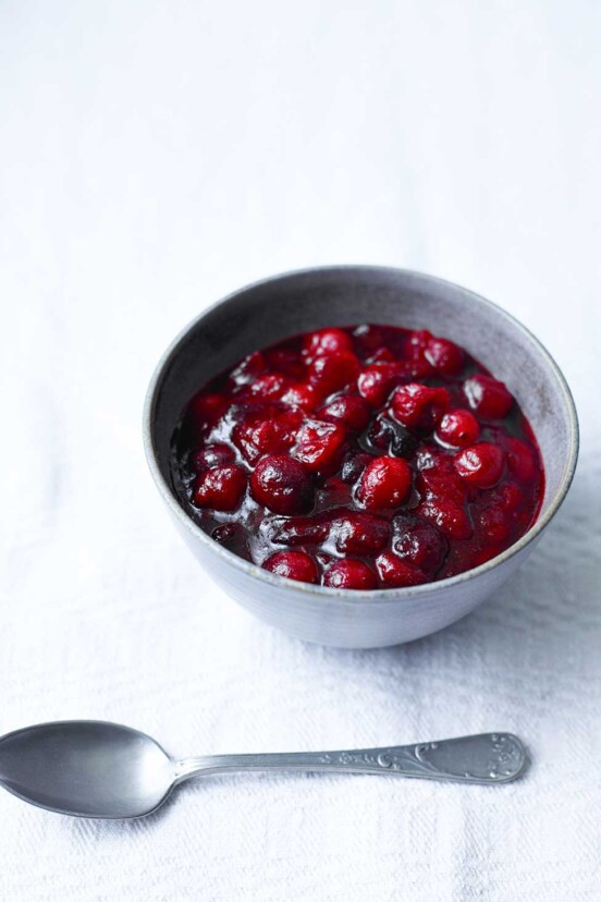 Spicy cranberry sauce in a grey pottery bowl on a white tablecloth, beside a serving spoon.