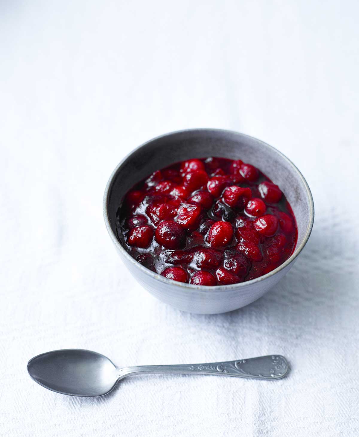 Spicy cranberry sauce in a grey pottery bowl on a white tablecloth, beside a serving spoon.