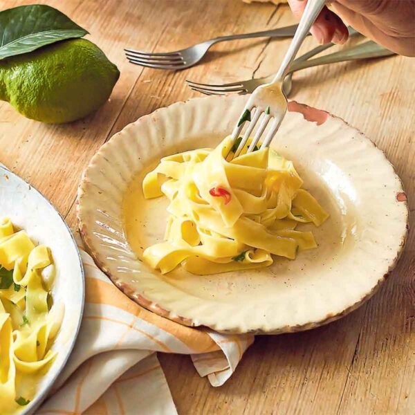 Tagliatelle with lemon on a large serving pate beside a smaller plate with noodles being twirled on a fork. Next to it, there are more forks and some lemons and a chunk of Parmesan.