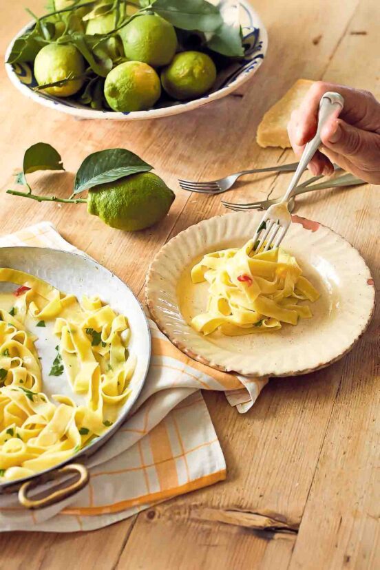 Tagliatelle with lemon on a large serving pate beside a smaller plate with noodles being twirled on a fork. Next to it, there are more forks and some lemons and a chunk of Parmesan.