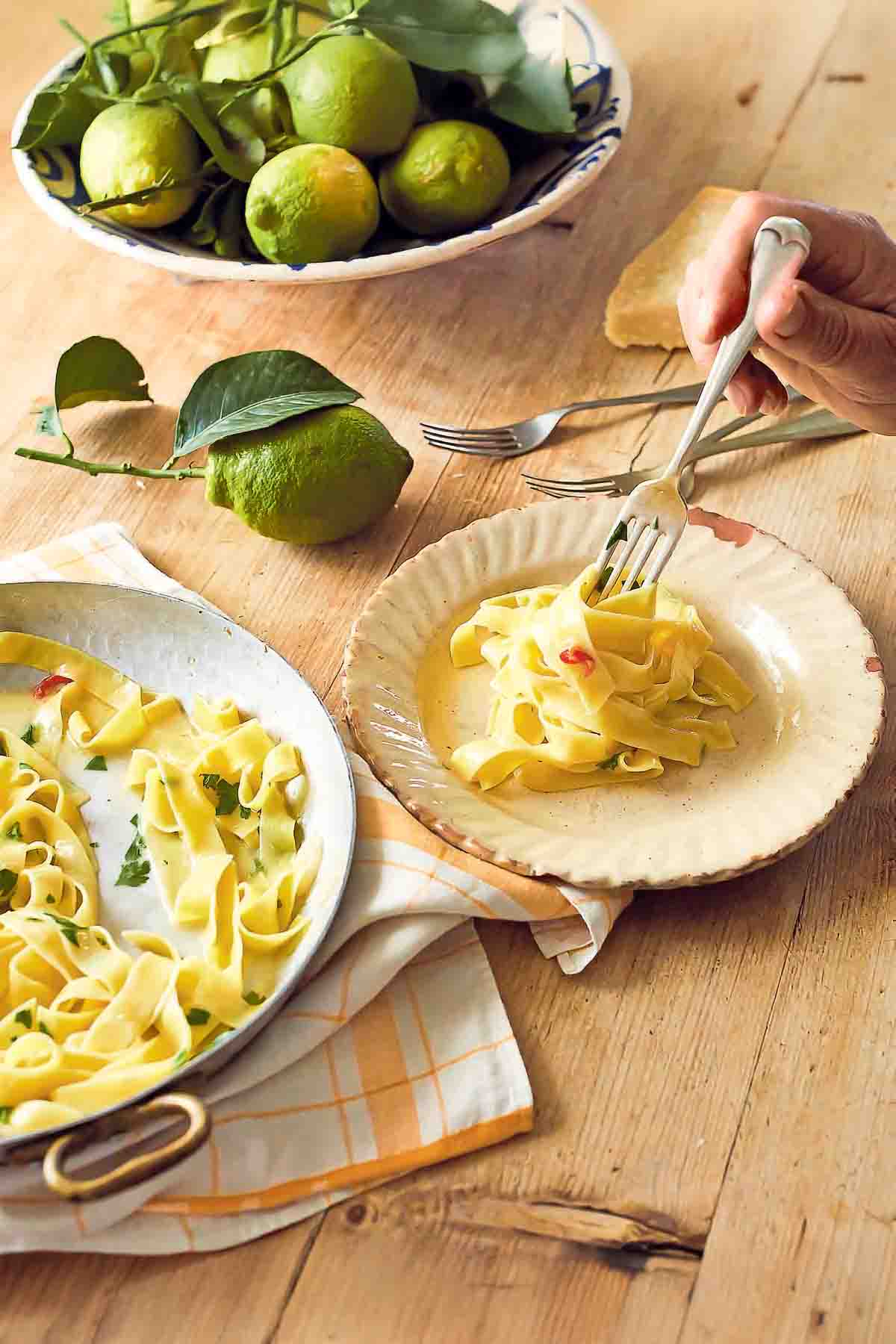 Tagliatelle with lemon on a large serving pate beside a smaller plate with noodles being twirled on a fork. Next to it, there are more forks and some lemons and a chunk of Parmesan.