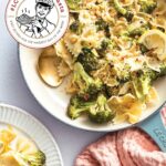Vegan lemon broccoli bow ties in a white enamel pan with a spoon and sliced lemons, beside a white plate full of pasta.