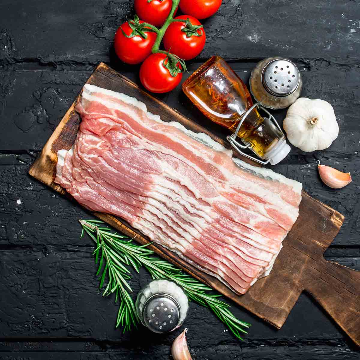 Strips of raw bacon on a cutting board with tomatoes, garlic, and rosemary