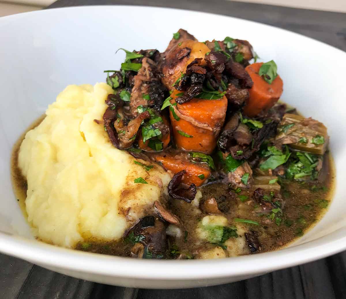 A white bowl with beef Bourguignon, filled with beef, carrots, and mushrooms, and mashed potatoes