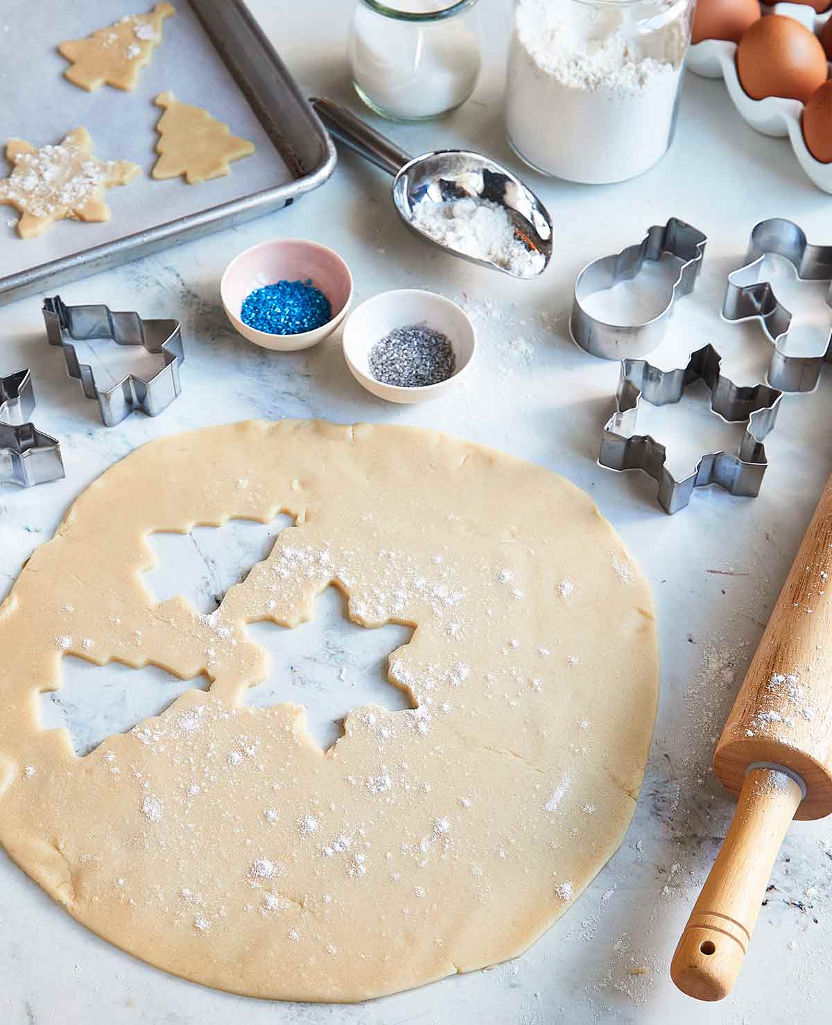 Christmas sugar cookies being rolled out and placed on a sheet pan, with cookie cutters, a rolling pin, sprinkles, and flour nearby.