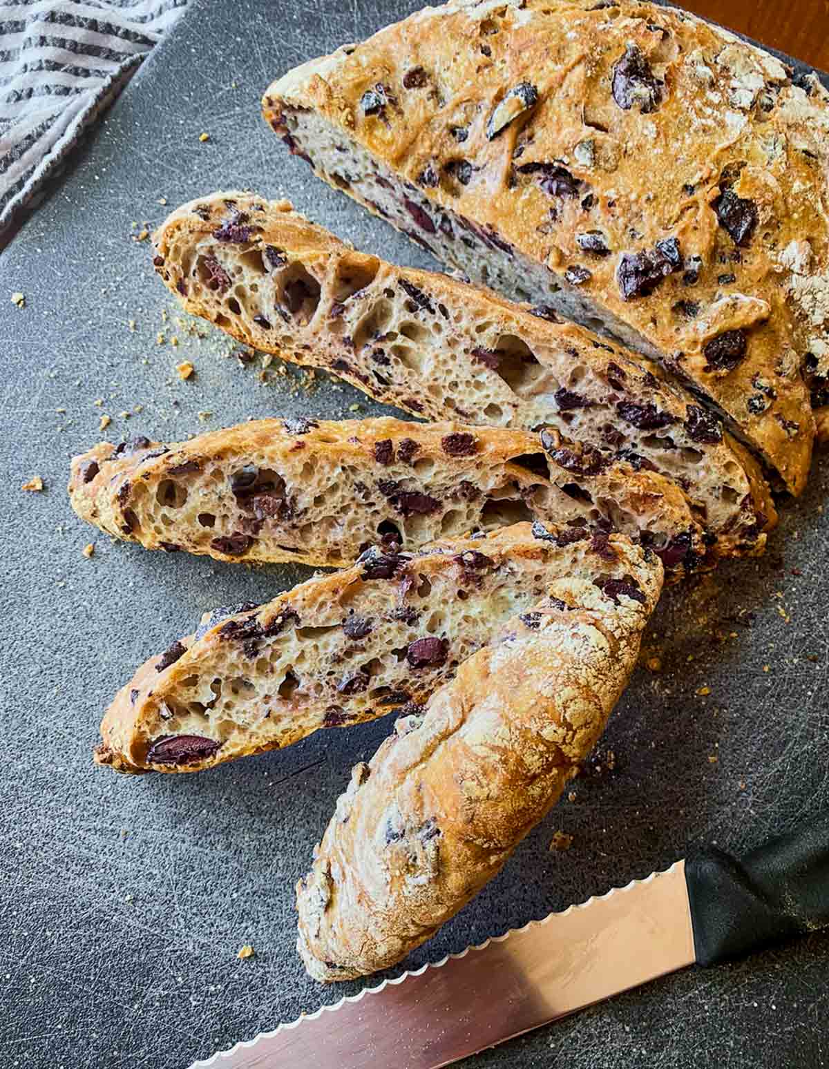 Jim Lahey no-knead olive bread with four slices cut, sitting on a blue cutting board with a napkin, a bread knife, and a pile of sliced black olives.