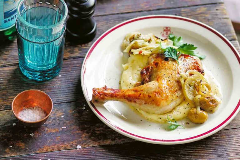 A plate with a leg and though of Mediterranean chicken with fennel and green olives sitting on a bed of mashed potatoes.