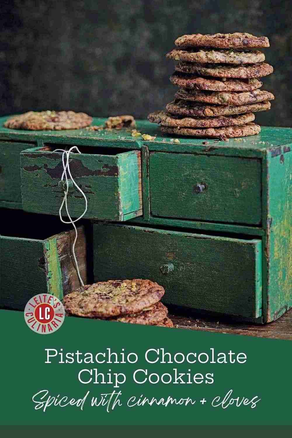 Several stacks of chocolate chip cookies studded with pistachio on a set of green wooden drawers.