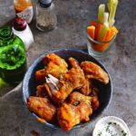 A bowl of buffalo chicken wings with a bottle of beer, a shot glass of raw vegetables, and a cup of dip on the side.