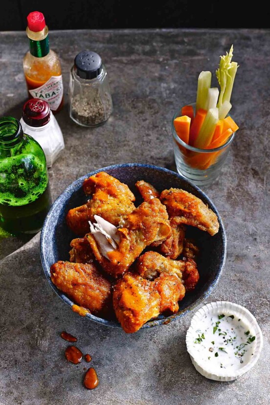 A bowl of buffalo chicken wings with a bottle of beer, a shot glass of raw vegetables, and a cup of dip on the side.