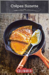 A small orange skillet with two crêpes Suzette and a spoon topped with citrus sauce.