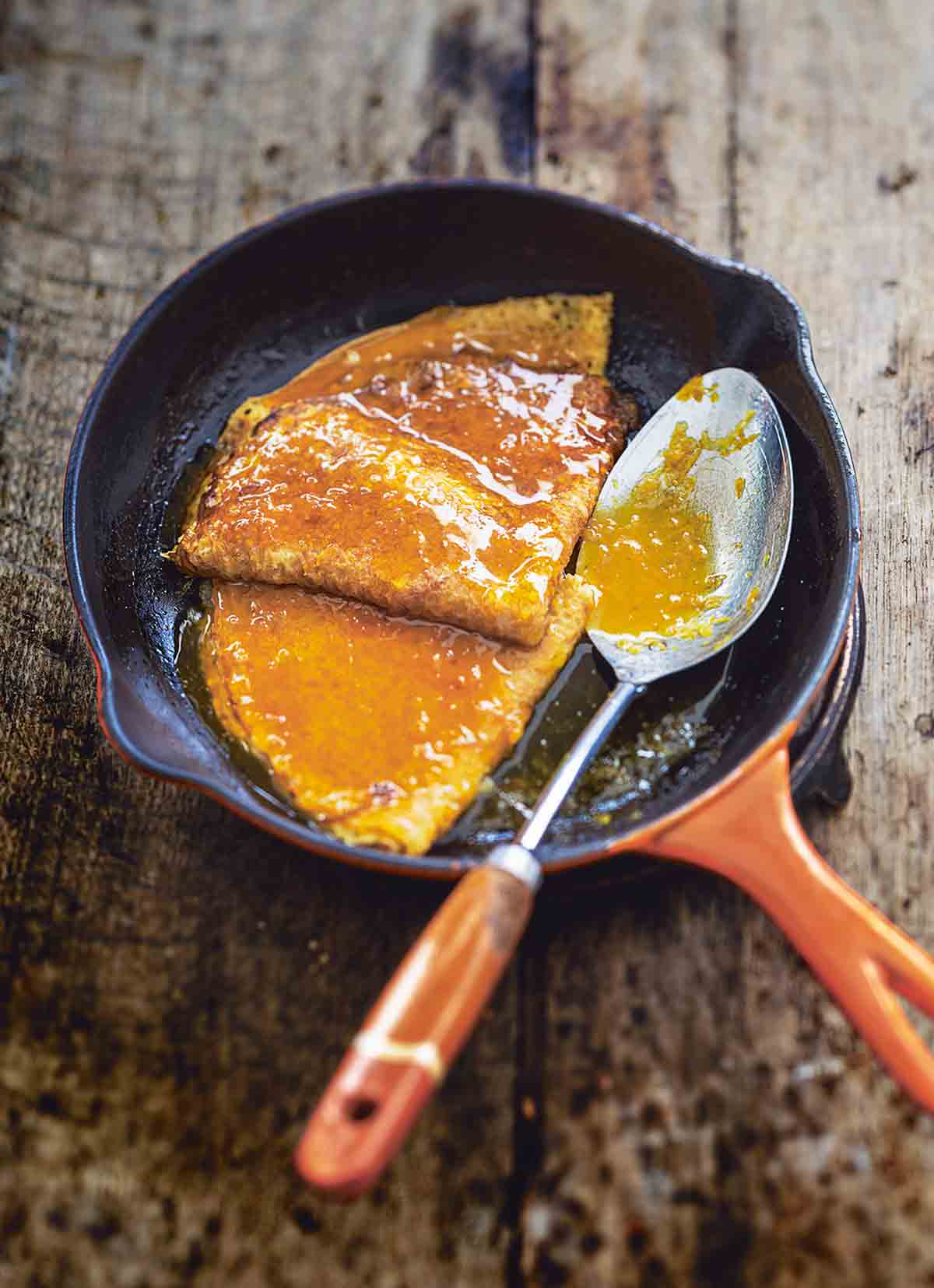 A small orange skillet with two crêpes Suzette and a spoon topped with citrus sauce.