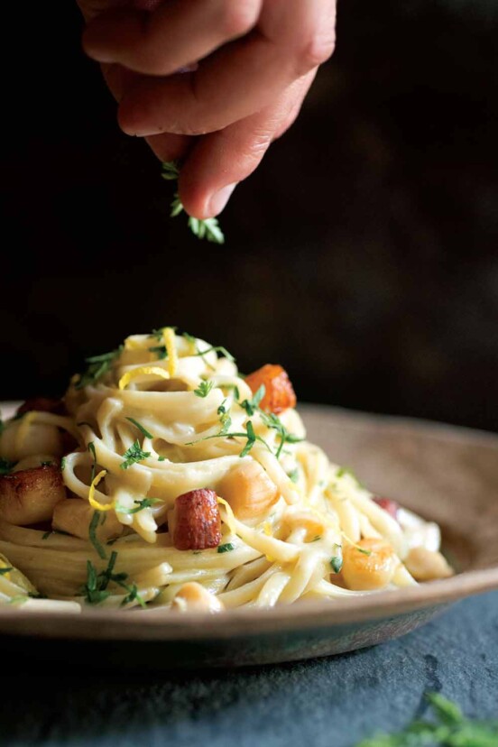 A tangle of fettuccine with scallops finished with lemon zest and chervil on a brown plate.