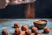 A dozen nutella truffles on a blue tabletop; a person's hand sifting cocoa over them