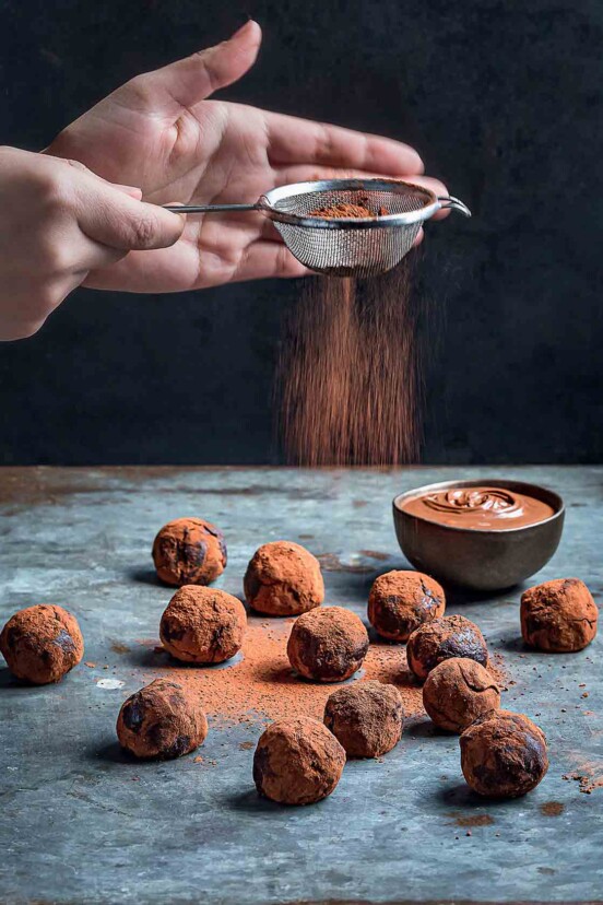 A dozen nutella truffles on a blue tabletop; a person's hand sifting cocoa over them