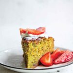A slice of orange, olive oil, and poppyseed cake with sliced strawberries on the side on a white plate