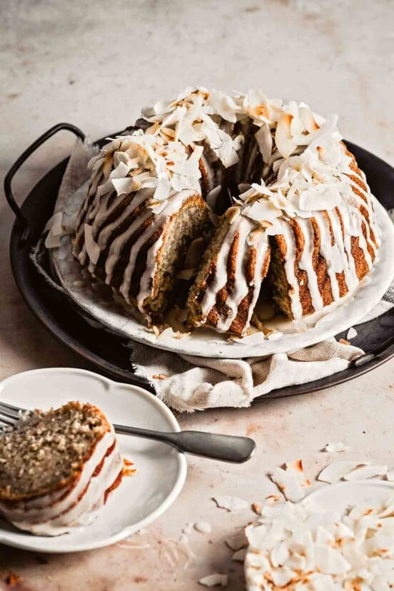A banana coconut rum bundt cake on a metal try with a slice cut from in on a plate nearby.