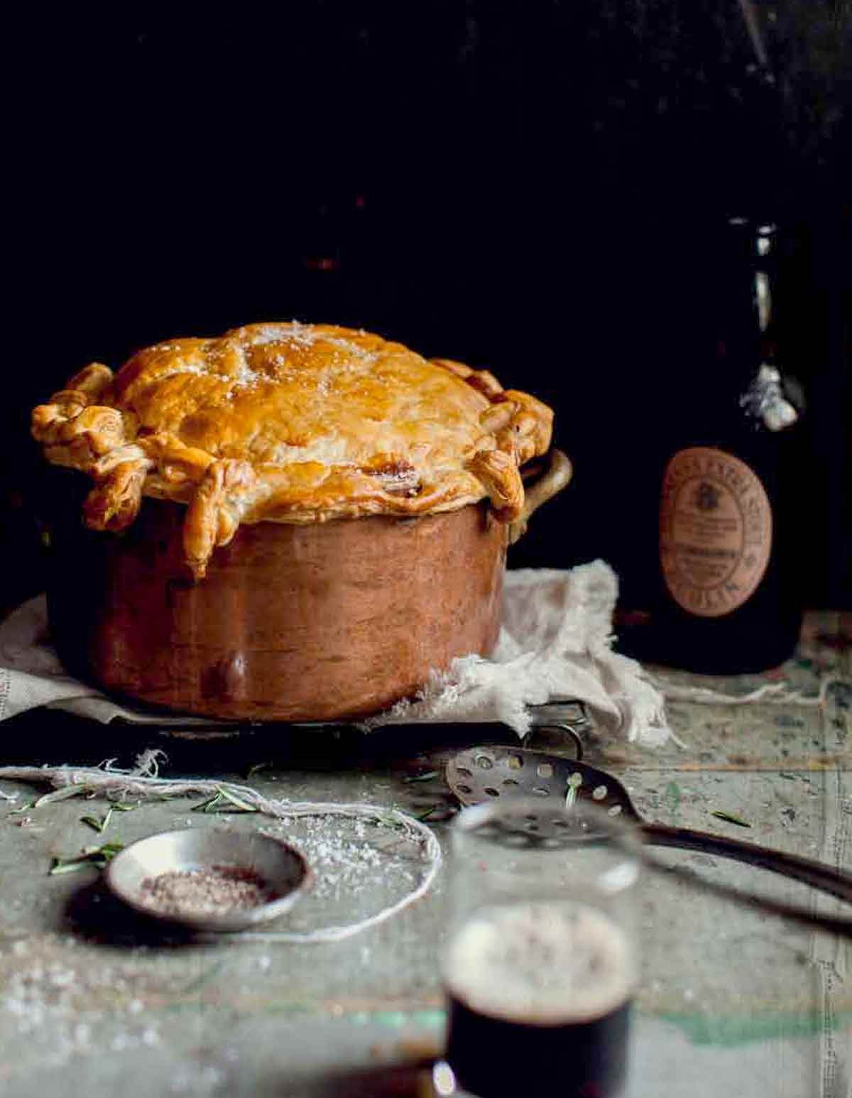 A copper pot filled with beef and Guinness cake with puff pastry on a towel on a grill with a bottle behind.