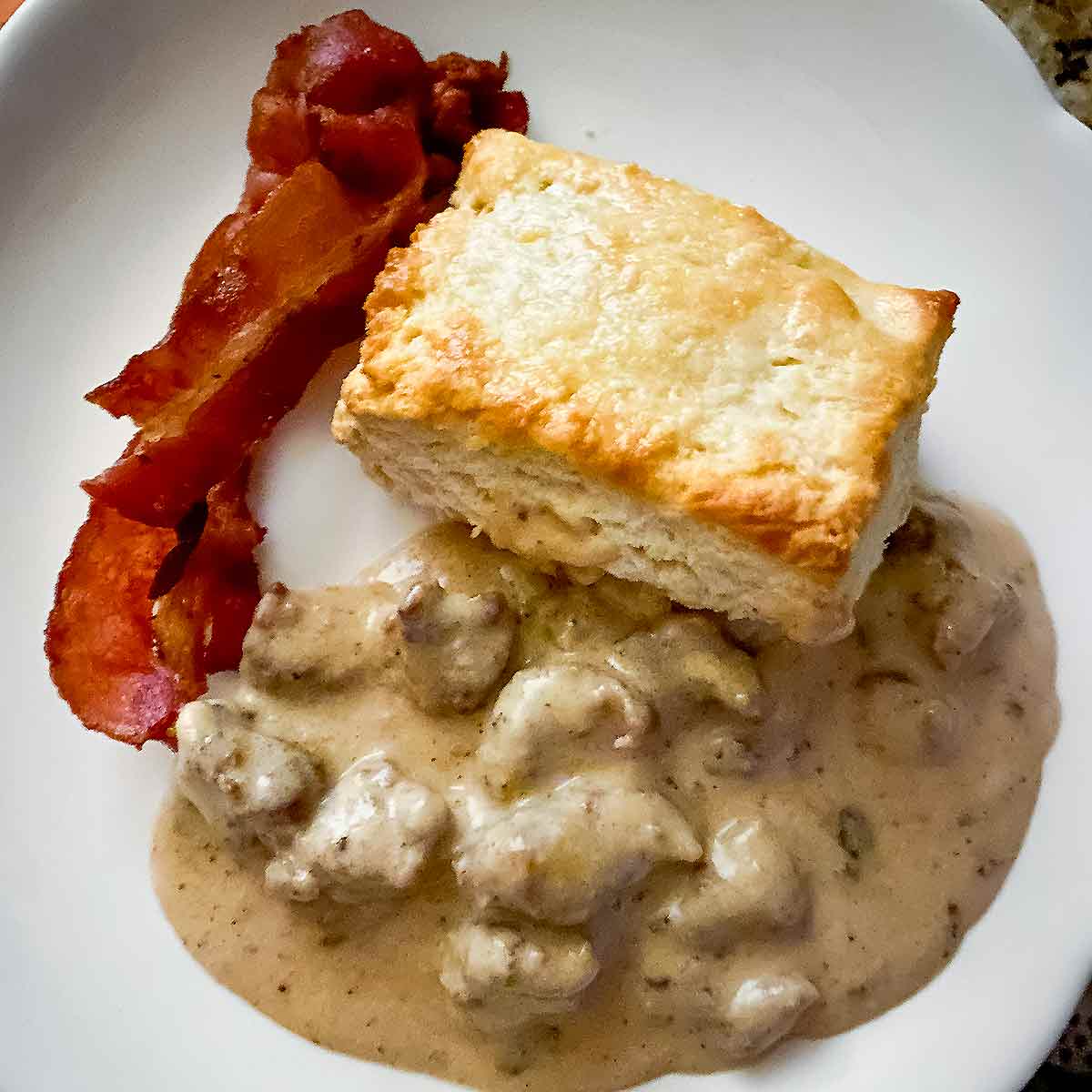 A white plate with a cat head biscuits and gravy and two strips of bacon.