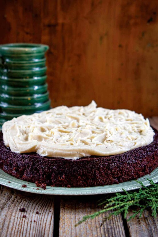 A chocolate stout cake topped with brown butter cream cheese frosting on a green plate with a green mug in the background.