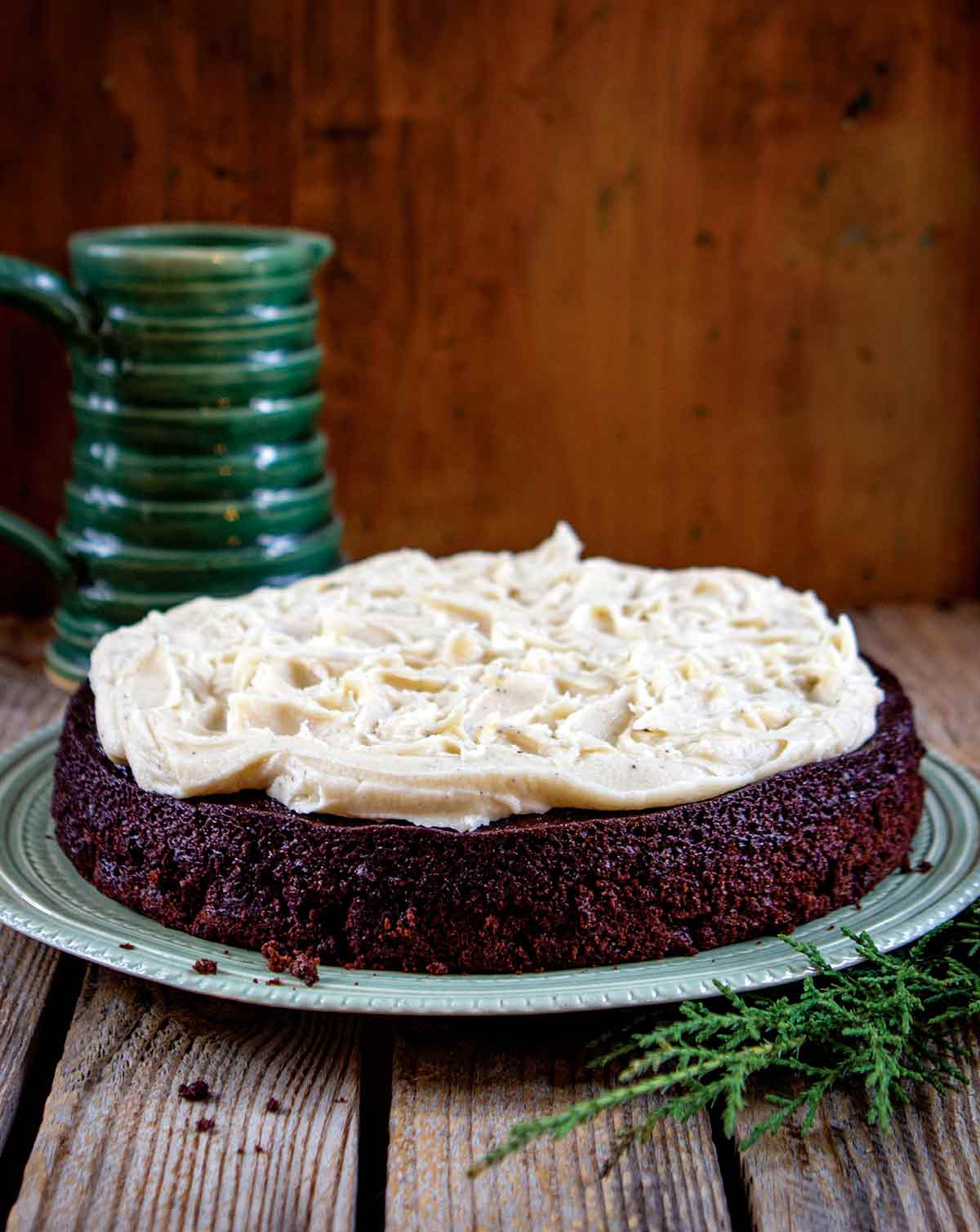 A chocolate stout cake with brown butter cream cheese frosting on a green plate with a green mug in the background.