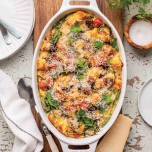 An oval casserole dish filled with easy breakfast strata on a wooden board with a wedge of Parmesan and a spoon on the side.