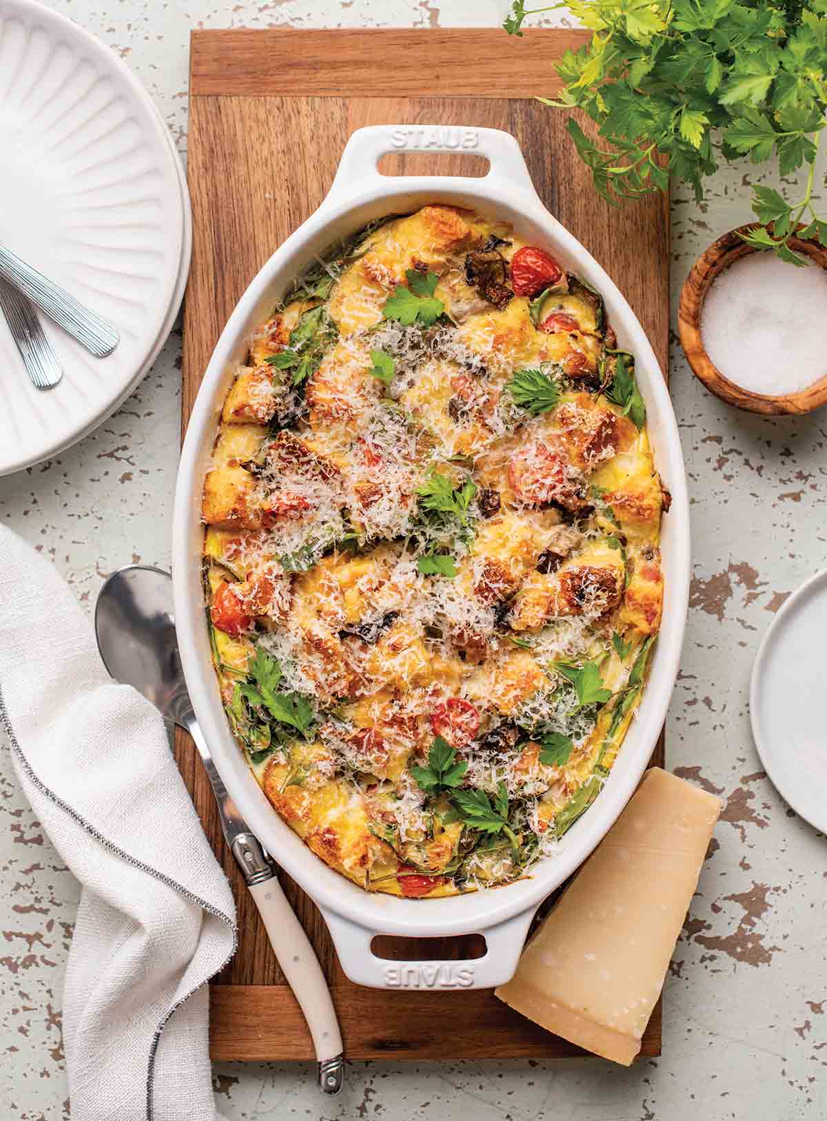 An oval casserole dish filled with easy breakfast strata on a wooden board with a wedge of Parmesan and a spoon on the side.