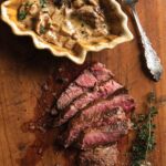 A sliced filet mignon with chanterelle marsala sauce in a bowl beside it on a wooden cutting board.