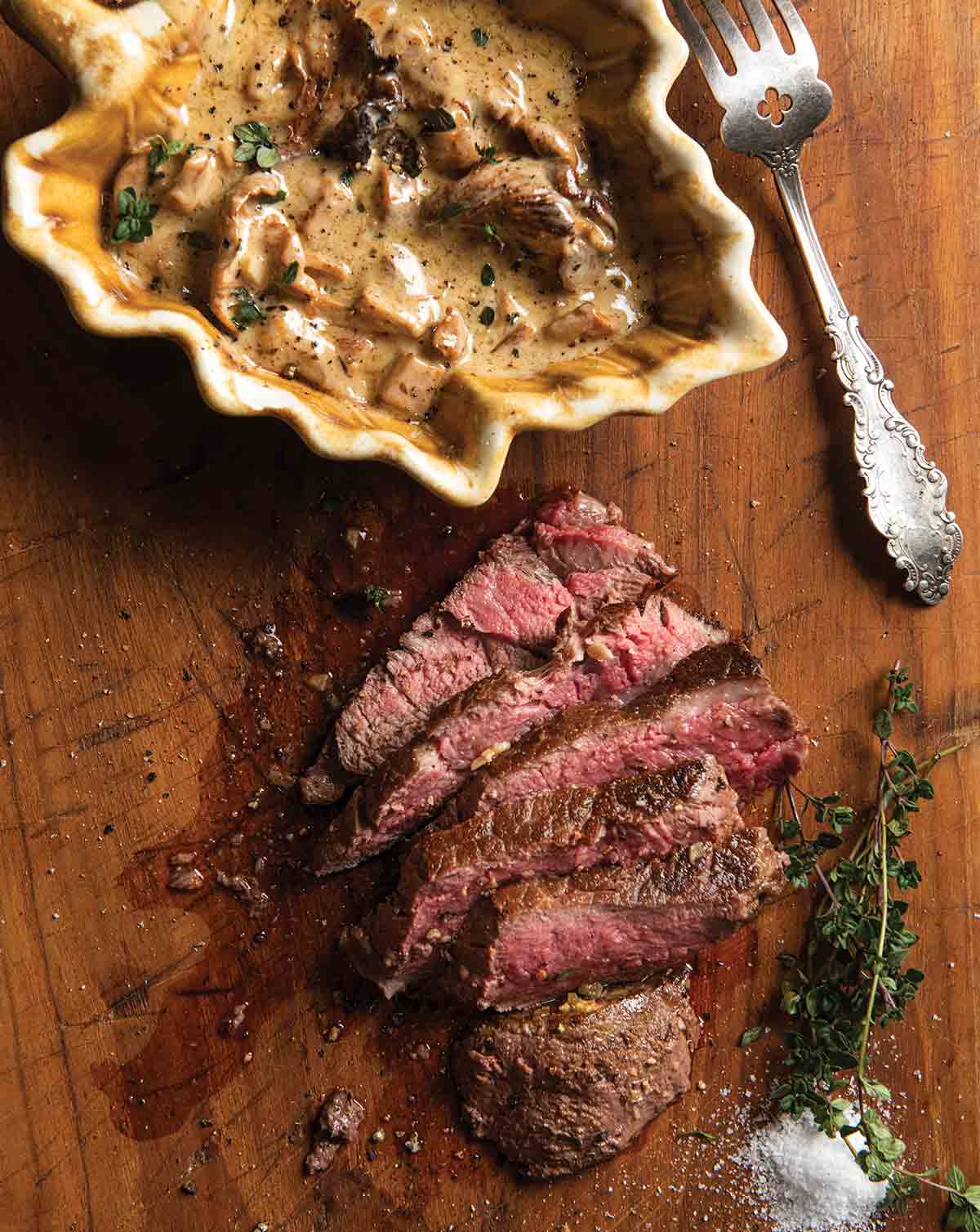A sliced filet mignon with chanterelle marsala sauce in a bowl beside it on a wooden cutting board.