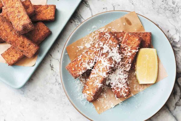 Four fried polenta sticks on a plate topped with Parmesan and a lemon wedge on the side.