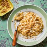 A fork resting in a tangle of gorgonzola and walnut linguine on a green and white plate with a bowl of toasted walnuts on the side.