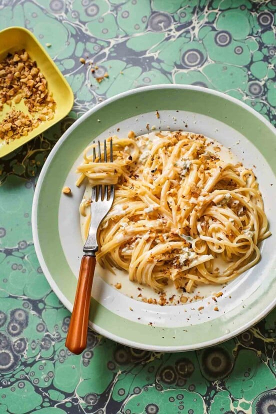 A fork resting in a tangle of gorgonzola and walnut linguine on a green and white plate with a bowl of toasted walnuts on the side.