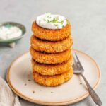 Five sweet potato patties stacked on a plate with a dollop of crème fraîche on top and a fork on the side.