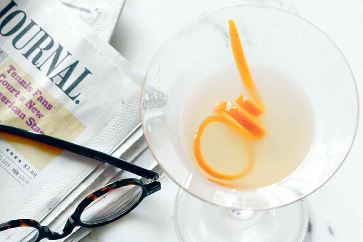 A breakfast martini with orange zest next to a plate with buttered toast and marmalade, a pair of reading glasses, and a newspaper.
