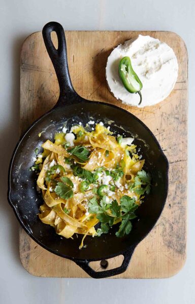 A cast-iron skillet filled with chilaquiles on a wooden cutting board with a block of queso fresco and half a jalapeno beside it.
