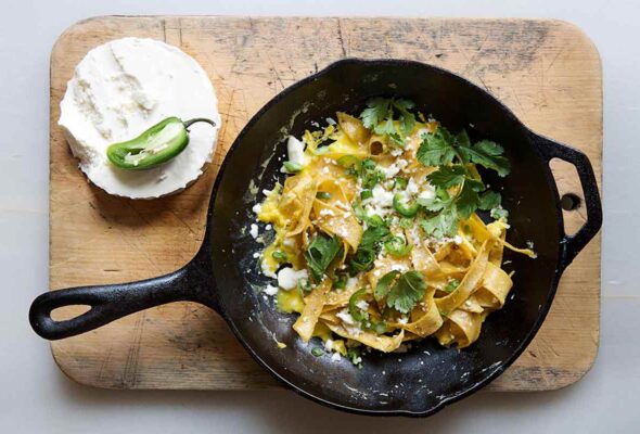 A cast-iron skillet filled with chilaquiles on a wooden cutting board with a block of queso fresco and half a jalapeno beside it.