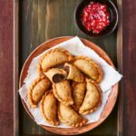 A pile of Chilean meat empanadas on a plate on a wooden tray with a dish of tomato relish on the side