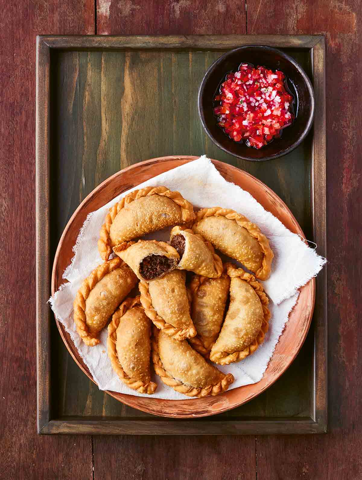A pile of Chilean meat empanadas on a plate on a wooden tray with a dish of tomato relish on the side