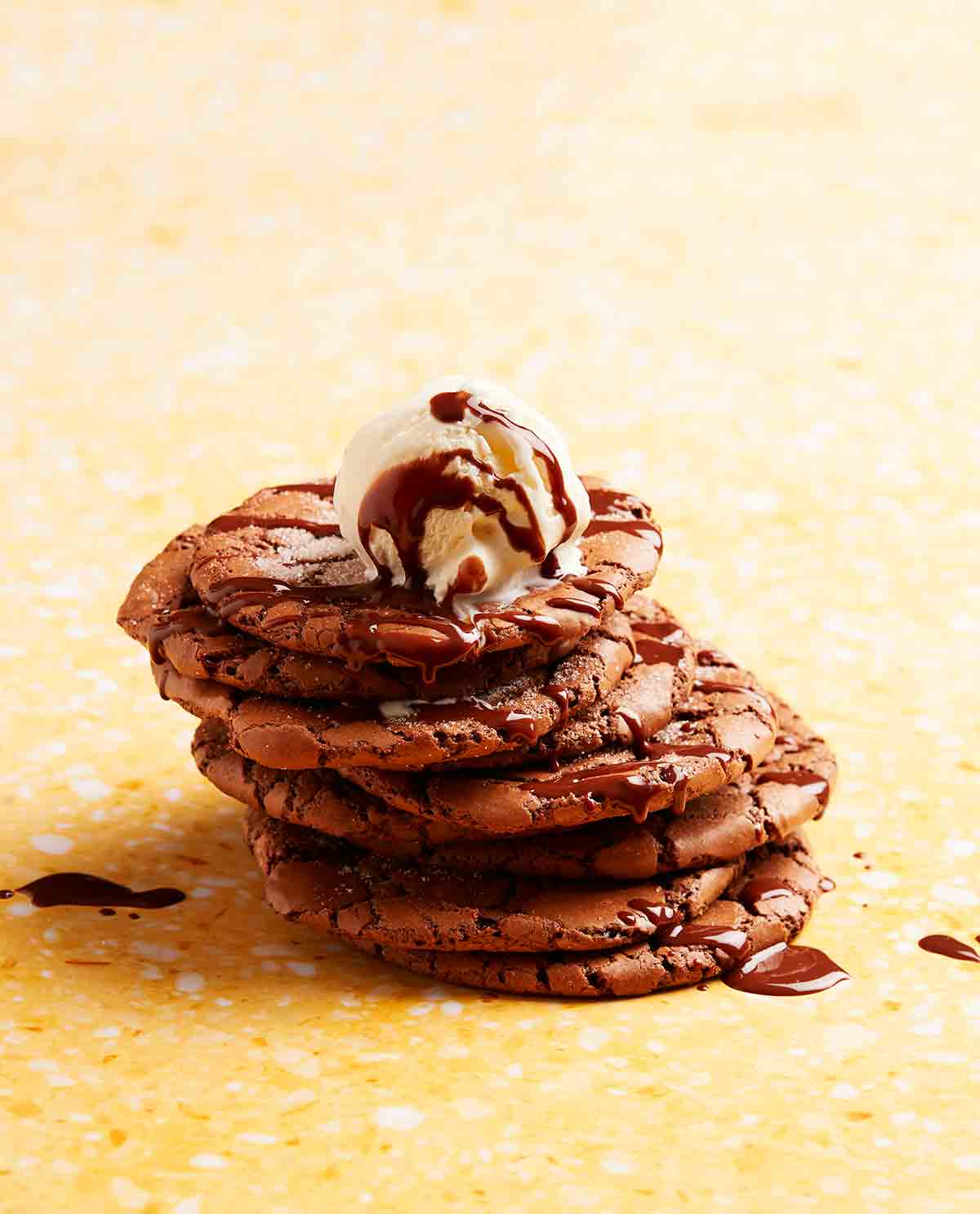 A stack of cracked brownie cookies with chocolate sauce and ice cream on top