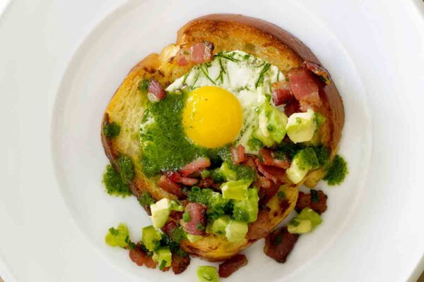 A slice of bread with a cooked egg in the center topped with ham, avocado salsa, and jalapeño oil, for a playful take on green eggs and ham.