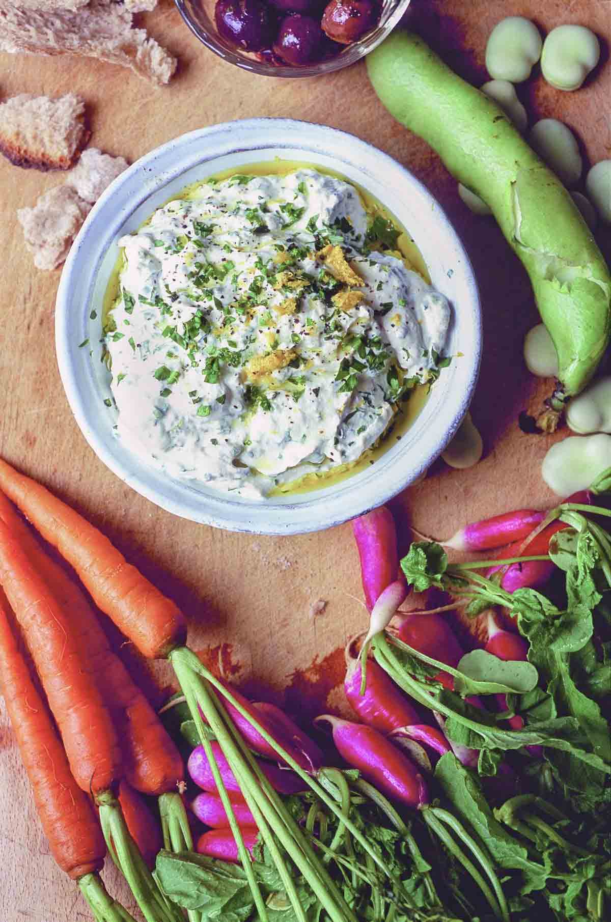 Herbed ricotta dip with a cutting board covered with carrots, radishes, fava beans