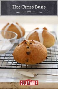 Three hot cross buns on a wire rack with a bowl of glaze and a spoon.