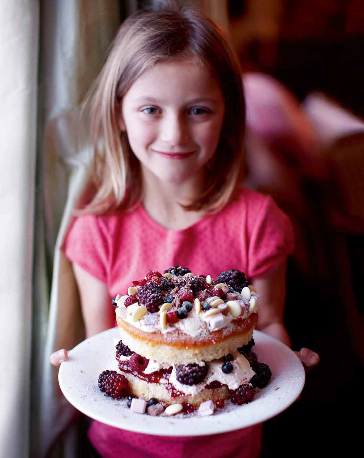 Ung flicka som håller en tvålagers isbärskaka fylld med hallon, blåbär, jordgubbar och vispgrädde
